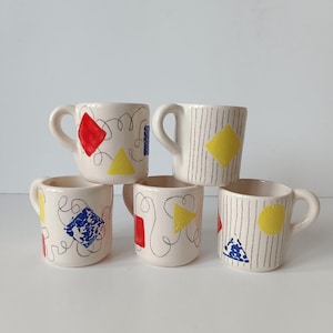 Può includere: Cinque piccole tazze in ceramica con manici. Ogni tazza è bianco sporco con un design unico con forme geometriche colorate come quadrati, triangoli e cerchi, insieme a linee nere ondulate. Alcune tazze hanno strisce verticali.