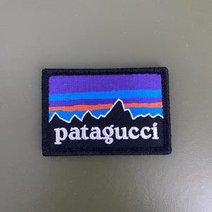 May include: Embroidered patch with a mountain range design and the word "patagucci" in white. The patch features a black border, a purple sky, and orange, blue, and white stripes. The patch is rectangular.