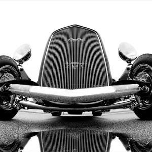 Könnte beinhalten: Schwarzweißfoto eines Oldtimer-Hot-Rods mit verchromtem Kühlergrill und Stoßstange. Das Auto steht auf einer schwarzen Asphaltfläche mit einer Spiegelung des Autos in einer Wasserpfütze.