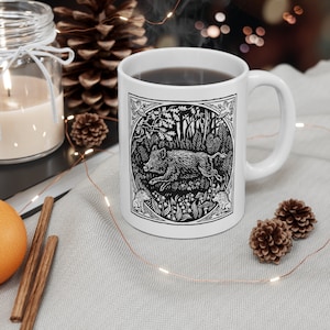 Può includere: Una tazza in ceramica bianca con un'illustrazione in bianco e nero di un cinghiale che corre attraverso una foresta. La tazza ha un manico nero ed è piena di caffè.