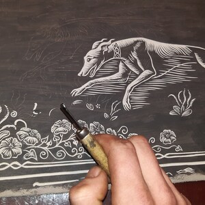 May include: A close-up of a woodblock print in progress. The print depicts a stylized dog running, a bird in flight, and floral designs. A hand holds a carving tool, working on the intricate details of the design.