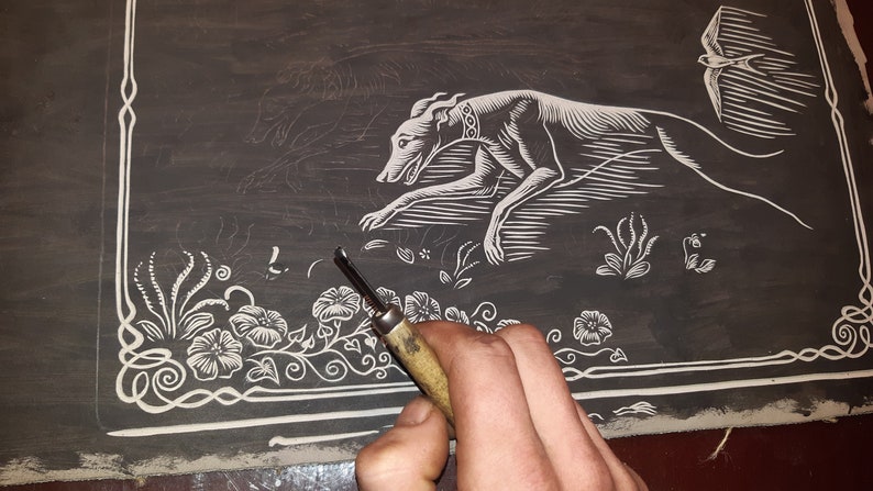 May include: A close-up of a woodblock print in progress. The print depicts a stylized dog running, a bird in flight, and floral designs. A hand holds a carving tool, working on the intricate details of the design.