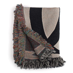 May include: A woven throw blanket with a black, white, and beige geometric pattern. The blanket has a fringe border.