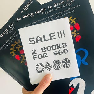 May include: Two books with dark covers, one titled "So Many Ways to Draw a Ghost." A white paper with "SALE!!! 2 BOOKS FOR $60" is held over the books. The books have illustrations of a swan, flowers, and gears.