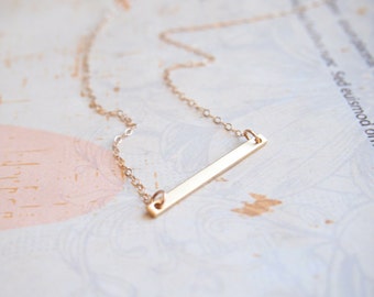 May include: A delicate gold chain necklace with a thin, horizontal bar pendant.