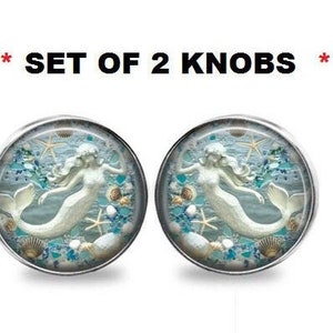 May include: Set of 2 round knobs with a mermaid design. Each knob features a white mermaid with flowing hair, surrounded by seashells, starfish, and blue accents. The knobs have a silver-colored metal rim.