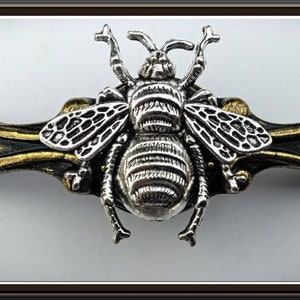 May include: A silver-colored bee ornament with detailed wings and body, mounted on a gold and bronze-colored decorative bar. The bee has intricate detailing, and the bar has a textured appearance. The design is likely for decorative purposes.