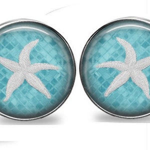 May include: A pair of round stud earrings featuring a white starfish design on a light blue background with a textured pattern. The earrings have a silver-colored metal border and a nautical theme.