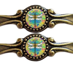 Op de afbeelding: Twee antieke bronzen ladehandgrepen. Elk heeft een centraal cirkelvormig ontwerp met een libellenillustratie op een kleurrijke achtergrond. De handgrepen hebben een decoratieve, gebogen handgreep.