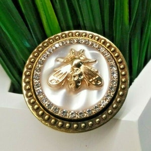 May include: A gold-toned bee-themed decorative button. The button features a detailed bee design in the center, surrounded by a ring of clear crystals and a textured gold border. The background is a pearlescent white.
