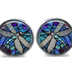 May include: A pair of round stud earrings with a dragonfly design. The earrings feature a stained-glass style with blue, teal, purple, and white colors. The dragonfly has detailed wings and a red body, set in a silver-colored metal.