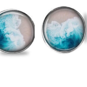 Könnte beinhalten: Ein Paar runde Ohrstecker mit silberfarbenem Rahmen. Jeder Ohrring zeigt eine Miniatur-Ozeanszene mit türkisfarbenen Wellen, die auf einen Sandstrand treffen. Das Design ist in klarem Harz eingeschlossen.