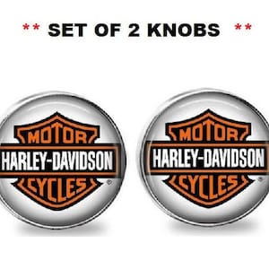 Può includere: Set di due pomelli rotondi con il logo Harley-Davidson. Il logo presenta un design arancione e nero con le parole "MOTOR HARLEY-DAVIDSON CYCLES" a forma di scudo. Il testo "SET OF 2 KNOBS" è in alto.