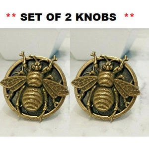 May include: Set of 2 decorative knobs. Each knob features a detailed, antique gold-colored bee design on a dark circular background. The bee is the focal point, with intricate wing and body details. The text "SET OF 2 KNOBS" is at the top.