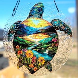 May include: A stained glass turtle suncatcher with a vibrant landscape scene. The design features a sunset over mountains, a river, and colorful flowers. The turtle's shell and flippers are detailed with a mosaic pattern.