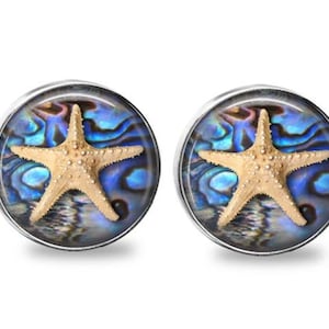 May include: A pair of round, silver-toned earrings. Each earring features a tan starfish design set against a swirling blue and iridescent background. The earrings are displayed on a white background.