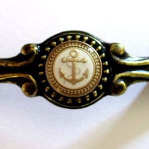 May include: Close-up of a decorative cabinet pull with a nautical theme. The pull features a central medallion with a white background and a gold anchor design. The surrounding frame is black and gold.