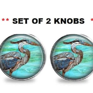 May include: Set of 2 round knobs featuring a detailed illustration of a blue heron against a teal and green background. The knobs have a silver-colored metal rim and are suitable for cabinets or drawers.