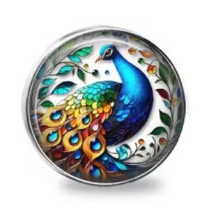 May include: A round, silver-toned button featuring a vibrant peacock design. The peacock has a blue head and neck, a colorful tail with yellow, orange, and blue accents, and is surrounded by green leaves and white floral elements.