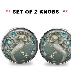 Puede incluir: Juego de dos pomos decorativos. Cada pomo presenta un borde metálico plateado y un diseño de sirena. La imagen muestra una sirena con una estrella de mar y conchas sobre un fondo turquesa.