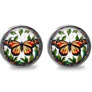 May include: A pair of round stud earrings featuring a monarch butterfly graphic with orange, black, and white wings, surrounded by green leaves on a white background, all encased in a silver-toned metal frame with a multi-colored border.