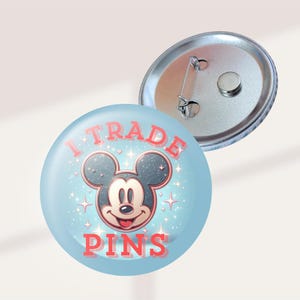 May include: A round button with a light blue background featuring a smiling Mickey Mouse face and the text "I TRADE PINS" in red. The button has a silver metal backing with a pin.