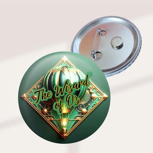 May include: A green button featuring a hot air balloon design with gold accents and the text "The Wizard of Oz." The button has a pin back and a metallic silver finish. The design is set against a dark green background.