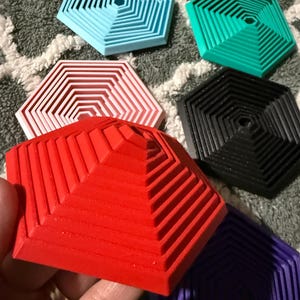May include: A collection of colorful, geometric coasters in various shades including red, blue, green, yellow, black, white, and purple. Each coaster has a hexagonal shape with a stepped, layered design. The coasters are made of a solid material.