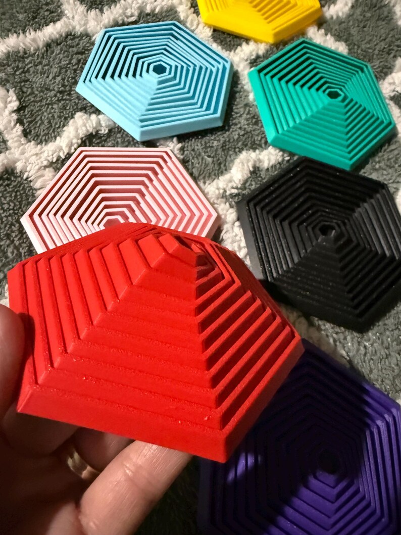May include: A collection of colorful, geometric coasters in various shades including red, blue, green, yellow, black, white, and purple. Each coaster has a hexagonal shape with a stepped, layered design. The coasters are made of a solid material.