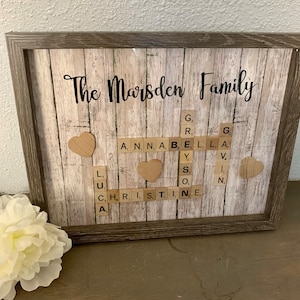 May include: A framed wooden sign with the text "The Marsden Family" in black lettering. The sign has a woodgrain background and features a scrabble tile design with the names "Anna", "Bella", "Gavin", and "Christine" spelled out. There are also small wooden hearts on the sign.