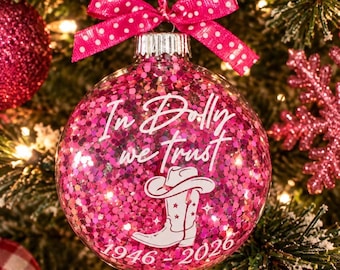 May include: A clear spherical Christmas ornament filled with bright pink glitter. The front features white text reading In Dolly we trust, a white cowboy boot graphic, and the years 1946-2026. A pink ribbon with white polka dots is tied at the top.