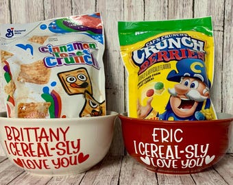 May include: Two white ceramic bowls with red hearts and the text "I cereal-sly love you" on them. One bowl says "Brittany" and the other says "Eric". The bowls are filled with cereal boxes. One box is Cinnamon Toast Crunch and the other is Cap'n Crunch's Crunch Berries.