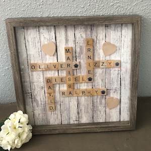 May include: A framed wooden sign with a white background and a wood grain pattern. The sign features a scrabble tile word art design with the names "Oliver", "Diesel", "Ernie", "M.A", "R.A.L.P.H", and "Z.Z.Y" spelled out in scrabble tiles. The names are surrounded by small wooden hearts.
