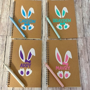 May include: Four brown spiral notebooks with bunny designs and personalized names. The notebooks have blue, purple, pink, and turquoise bunny ears and bows. Each notebook comes with a pen.