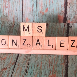May include: A wooden name sign with the name "GONZALEZ" spelled out in Scrabble tiles. The tiles are arranged on a wooden base with a mermaid figurine on the left and a Flounder and Sebastian figurine on the right.