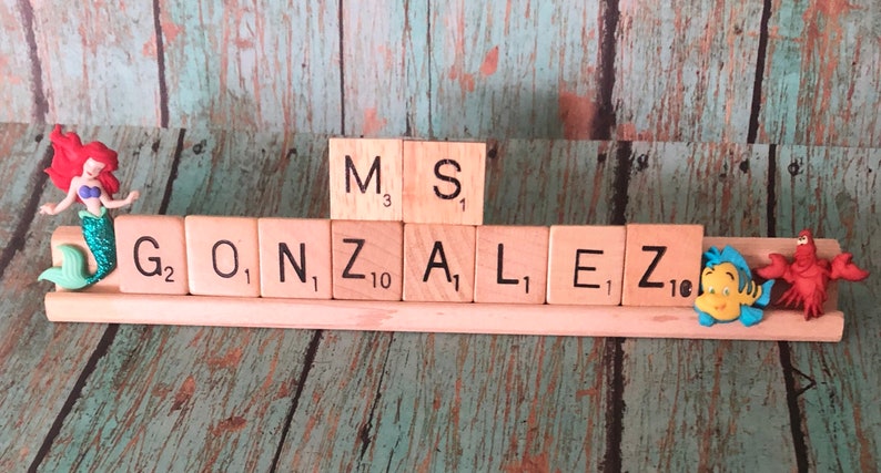 May include: A wooden name sign with the name "GONZALEZ" spelled out in Scrabble tiles. The tiles are arranged on a wooden base with a mermaid figurine on the left and a Flounder and Sebastian figurine on the right.