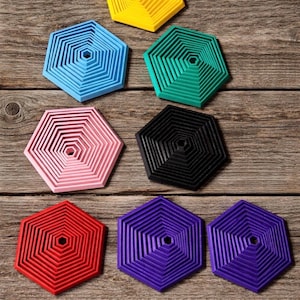 May include: A collection of colorful, hexagonal coasters in various shades, including yellow, blue, green, pink, black, red, and purple. The coasters have a layered design and are arranged on a wooden surface.