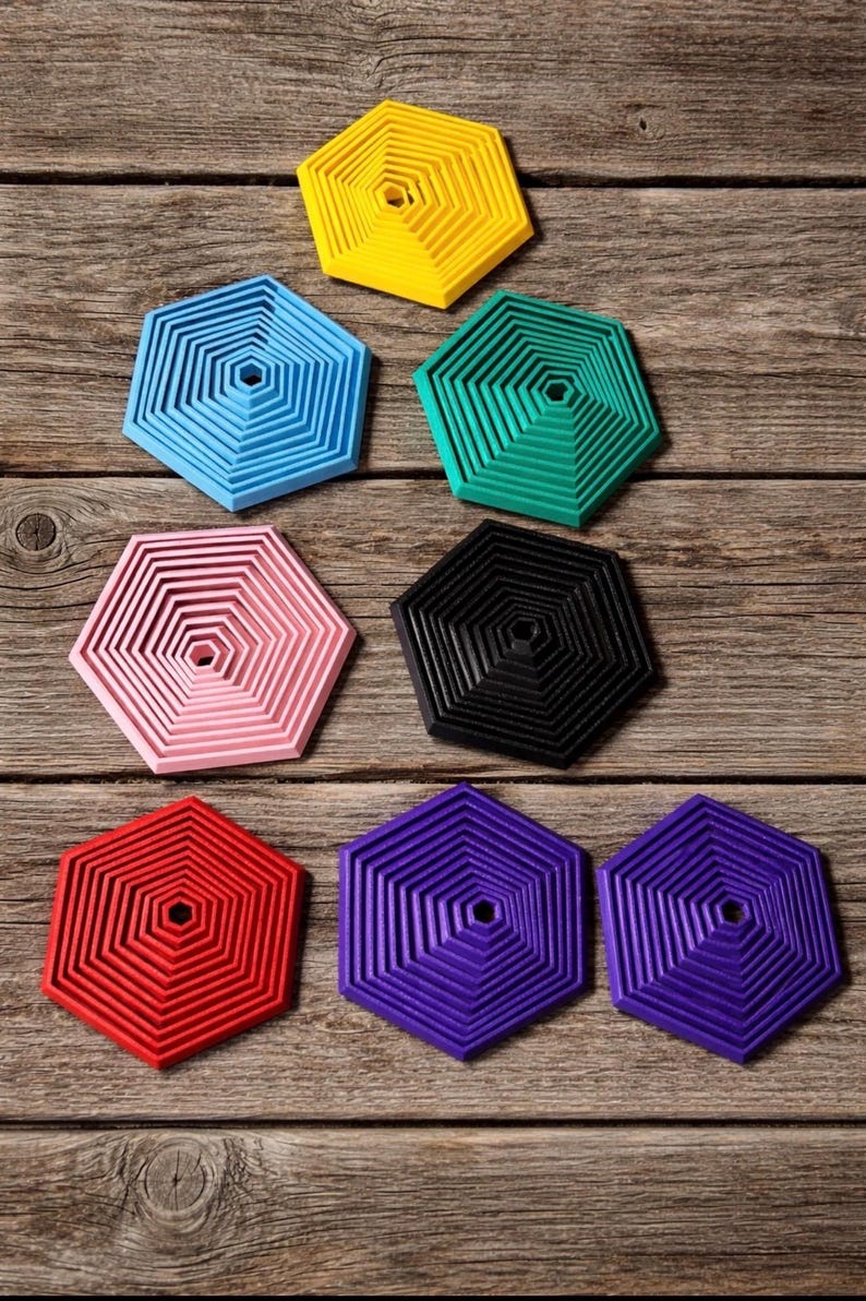 May include: A collection of colorful, hexagonal coasters in various shades, including yellow, blue, green, pink, black, red, and purple. The coasters have a layered design and are arranged on a wooden surface.