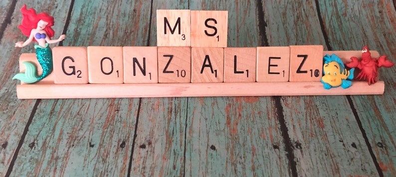 May include: A wooden name sign with the name "GONZALEZ" spelled out in Scrabble tiles. The tiles are arranged on a wooden base with a mermaid figurine on the left and a cartoon fish and lobster figurine on the right.