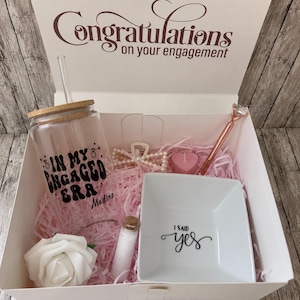 May include: A white gift box with the text "Congratulations on your engagement." Inside are a glass tumbler, a small dish that says "I said yes," a pen, a candle, a rose, and other items, all on pink paper.