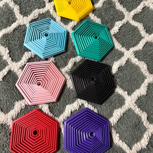 May include: A collection of seven hexagonal coasters in various colors: yellow, light blue, teal, pink, black, red, and purple. Each coaster features a layered, geometric design with a central hole. The coasters are arranged on a gray and white patterned surface.