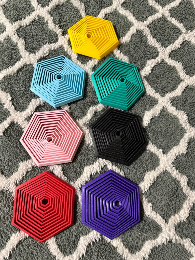 May include: A collection of seven hexagonal coasters in various colors: yellow, light blue, teal, pink, black, red, and purple. Each coaster features a layered, geometric design with a central hole. The coasters are arranged on a gray and white patterned surface.