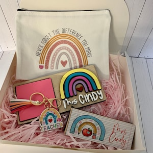 Puede incluir: Una caja de regalo blanca llena de papel de seda rosa y regalos de agradecimiento para maestros. Los regalos incluyen una bolsa de mano con temática de arcoíris con el texto "Nunca olvides la diferencia que haces", un letrero de arcoíris de madera con el texto "Sra. Cindy", un bloc de notas rosa, un llavero con un arcoíris y la palabra "Maestro", y un letrero de madera con un diseño de regla y manzana y el texto "Sra. Cindy".
