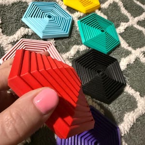 May include: A collection of colorful, geometric coasters in various shades including red, yellow, blue, green, black, and purple. The coasters have a layered, hexagonal design. One red coaster is being held, with others scattered on a patterned surface.