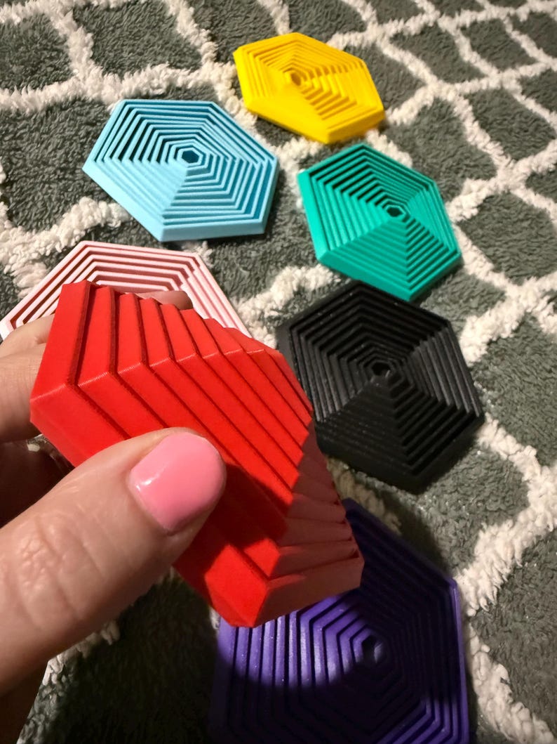 May include: A collection of colorful, geometric coasters in various shades including red, yellow, blue, green, black, and purple. The coasters have a layered, hexagonal design. One red coaster is being held, with others scattered on a patterned surface.