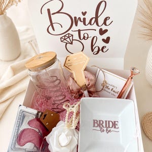 May include: A white gift box labelled Bride to be containing a glass tumbler with a bamboo lid and straw, a pink scrunchie on a card, a rose gold pen with a diamond topper, a white square trinket dish, a pearl hair clip, and pink eye masks.