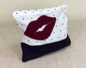 May include: A white zippered pouch with black polka dots and a red felt lip design. The pouch has a black fabric bottom.