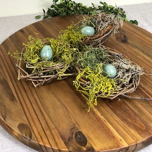 May include: Three twig nests with green moss and blue speckled eggs on a round wooden serving tray.