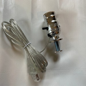 May include: A silver light socket with a clear cord and a plug. The socket has a screw-on base and a threaded adjustment knob. The cord is coiled and the plug is standard.