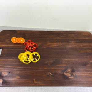 May include: A large, rectangular wooden cutting board with a handle. The board is dark brown and has a natural wood grain. There are slices of orange and yellow bell pepper on the board.